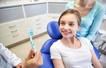 A young girl in a dentist's chair being educated on oral health by a dentist showing how to properly use a toothbrush.