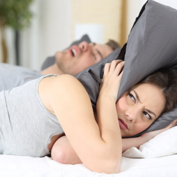 the woman covers her head with a pillow while the man sleeping next to her is snoring