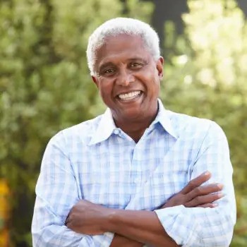 A man with gray hair smiles while standing outdoors with his arms crossed, wearing a light blue checkered shirt. The background is blurred greenery.