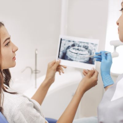 A dentist wearing blue gloves discusses a dental X-ray with a seated female patient in a dental clinic.