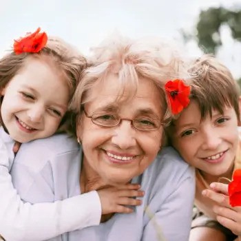 An elderly woman is smiling with two children, one on each side. They all have red flowers in their hair and are outdoors.