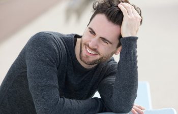 A person with dark hair and a beard leans on a surface, smiling. They are wearing a dark long-sleeve shirt.