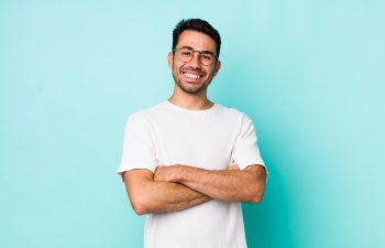 A smiling young man with glasses and a beard standing with his arms crossed against a light blue background.