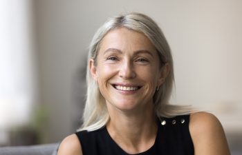 A close-up portrait of a smiling middle-aged woman with light blonde hair, wearing a black sleeveless top, looking directly at the camera. The background is blurred, showing a light, modern indoor setting.