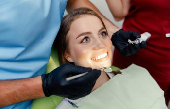 A smiling young woman lies in a dental chair looking up, while a dentist wearing black gloves examines her illuminated teeth with a dental mirror and a drill.