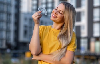 A happy young woman with long blonde hair and freckles smiles with her eyes closed as she holds up a transparent dental aligner. She is wearing a yellow t-shirt and is standing outdoors in a city, with modern apartment buildings blurred in the background.