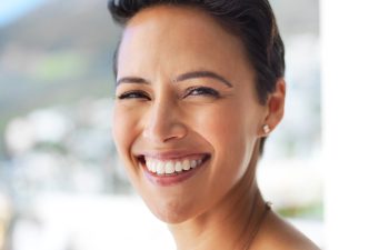 A close-up portrait of a smiling person with a short, dark pixie cut, looking at the camera with a bright, happy expression against a blurred, brightly lit background.