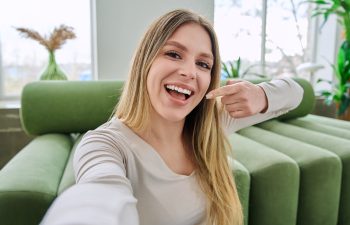 A cheerful woman with long blonde hair taking a selfie on a green sofa, smiling broadly and pointing at her white teeth.
