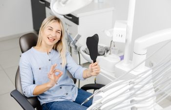 A blonde woman sitting in a dental chair, holding a tooth-shaped mirror, winking, and showing an OK sign with her hand.