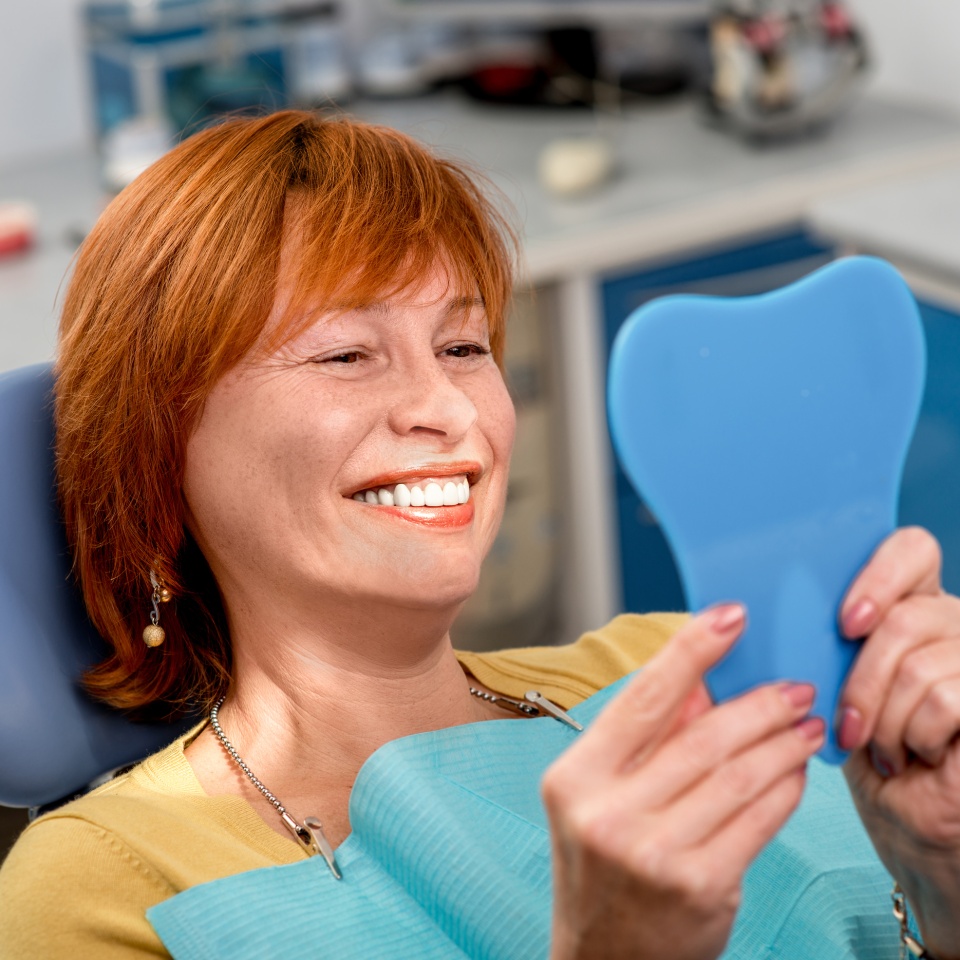 Smiling senior woman with new dental implants sitting in the dental office and looking at the mirror