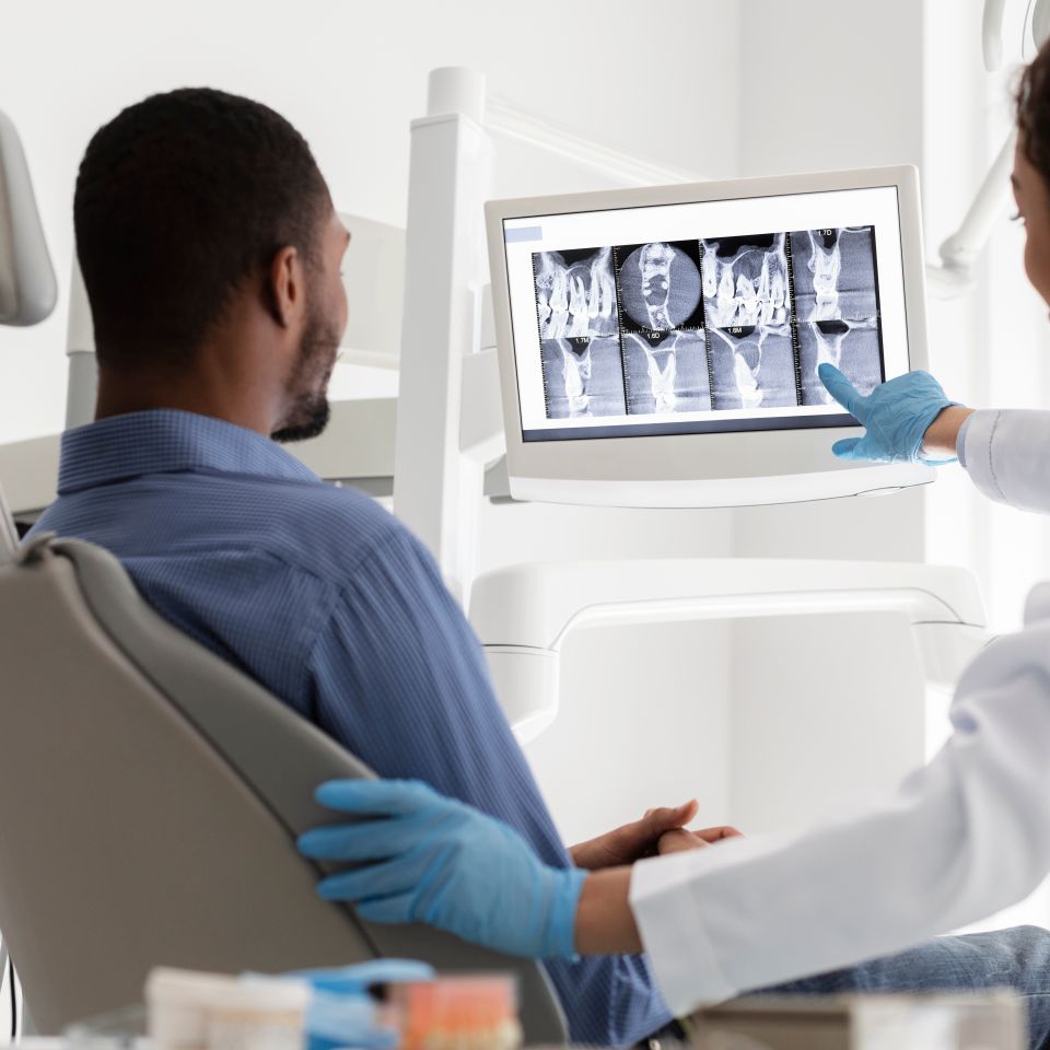 A dentist wearing gloves shows a dental X-ray image on a monitor to a patient sitting in a dental chair.