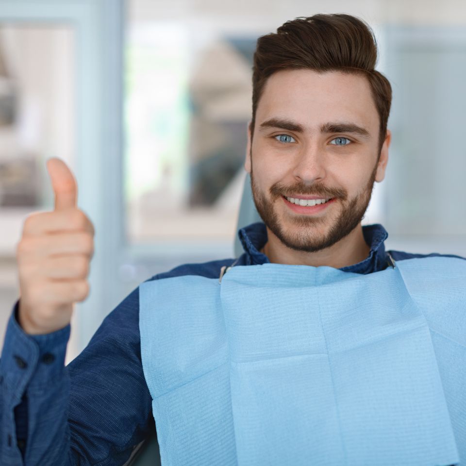 A man sitting in a dental chair wearing a blue dental bib gives a thumbs up and smiles at the camera.
