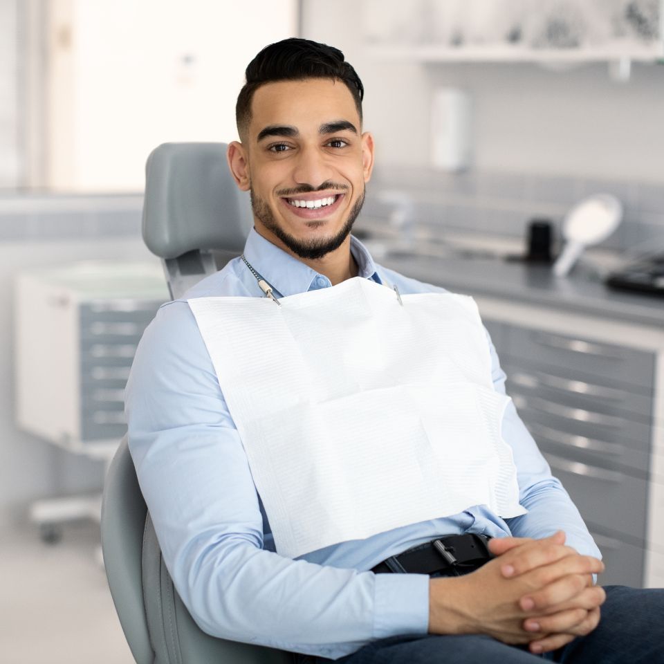 A man sits in a dentist's chair, smiling, wearing a light blue shirt and a dental bib, in a modern dental clinic.