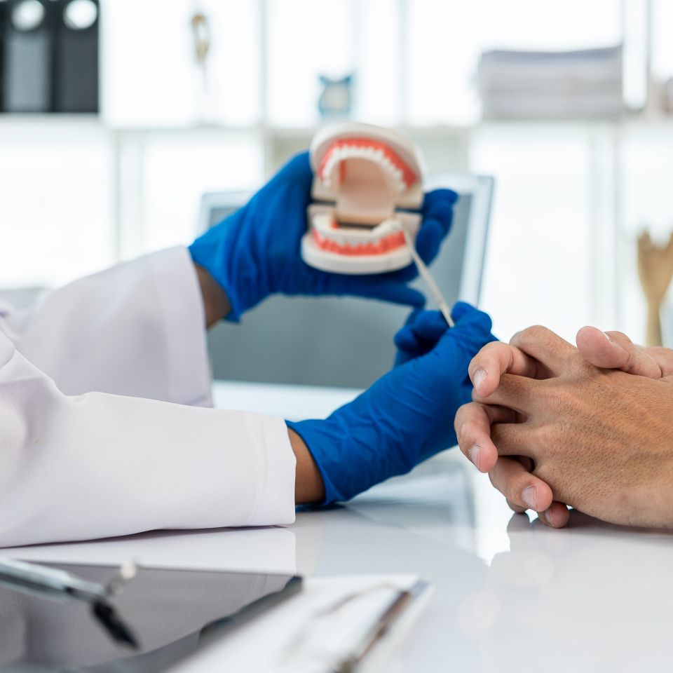 A dentist wearing blue gloves uses a dental model and tool to explain oral care to a patient seated across a desk.