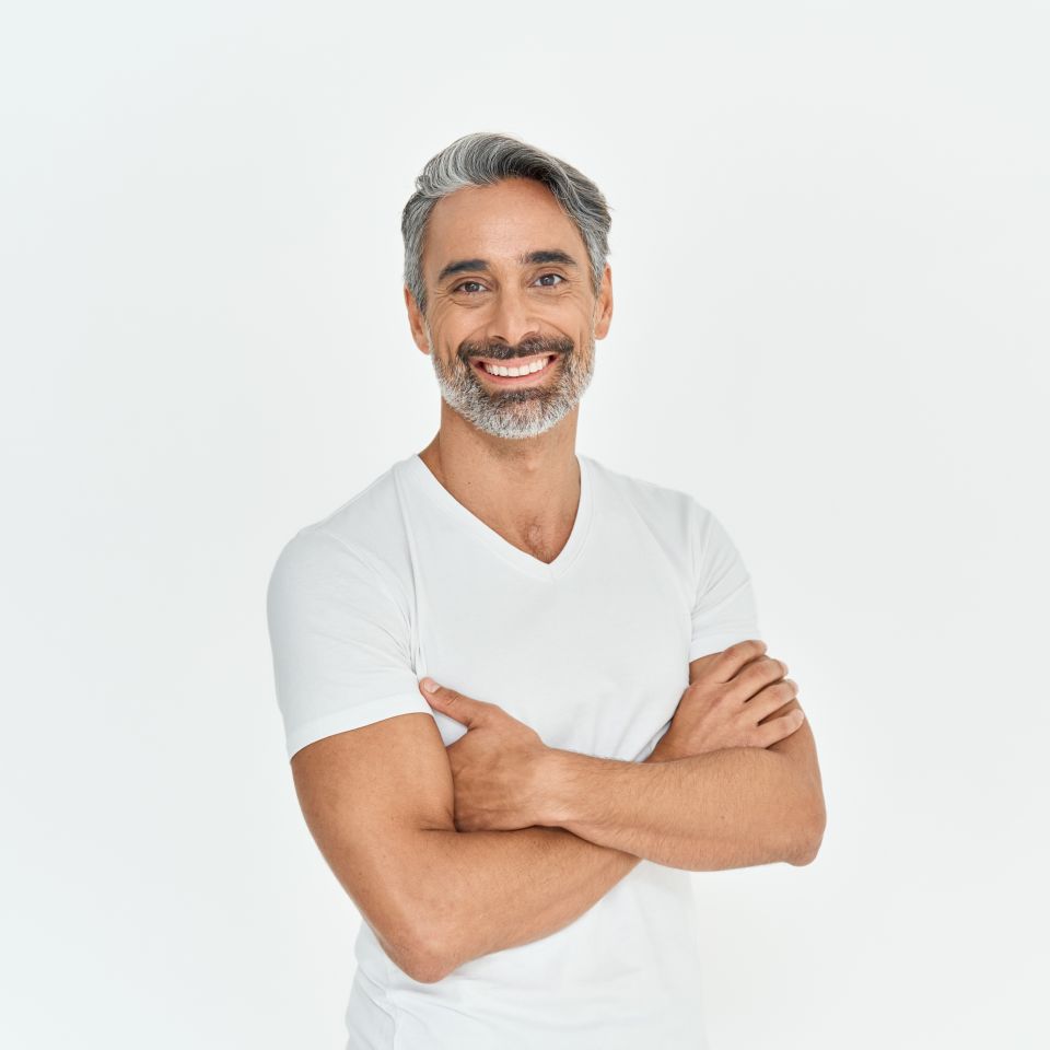 A man with gray hair and a beard, wearing a white t-shirt, stands against a plain white background with his arms crossed and smiling.